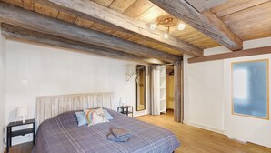 Ein Schlafzimmer mit Holzboden, einem Bett mit kariertem Überwurf, einem Nachttisch mit Lampe und einer Tür zu einem anderen Raum.