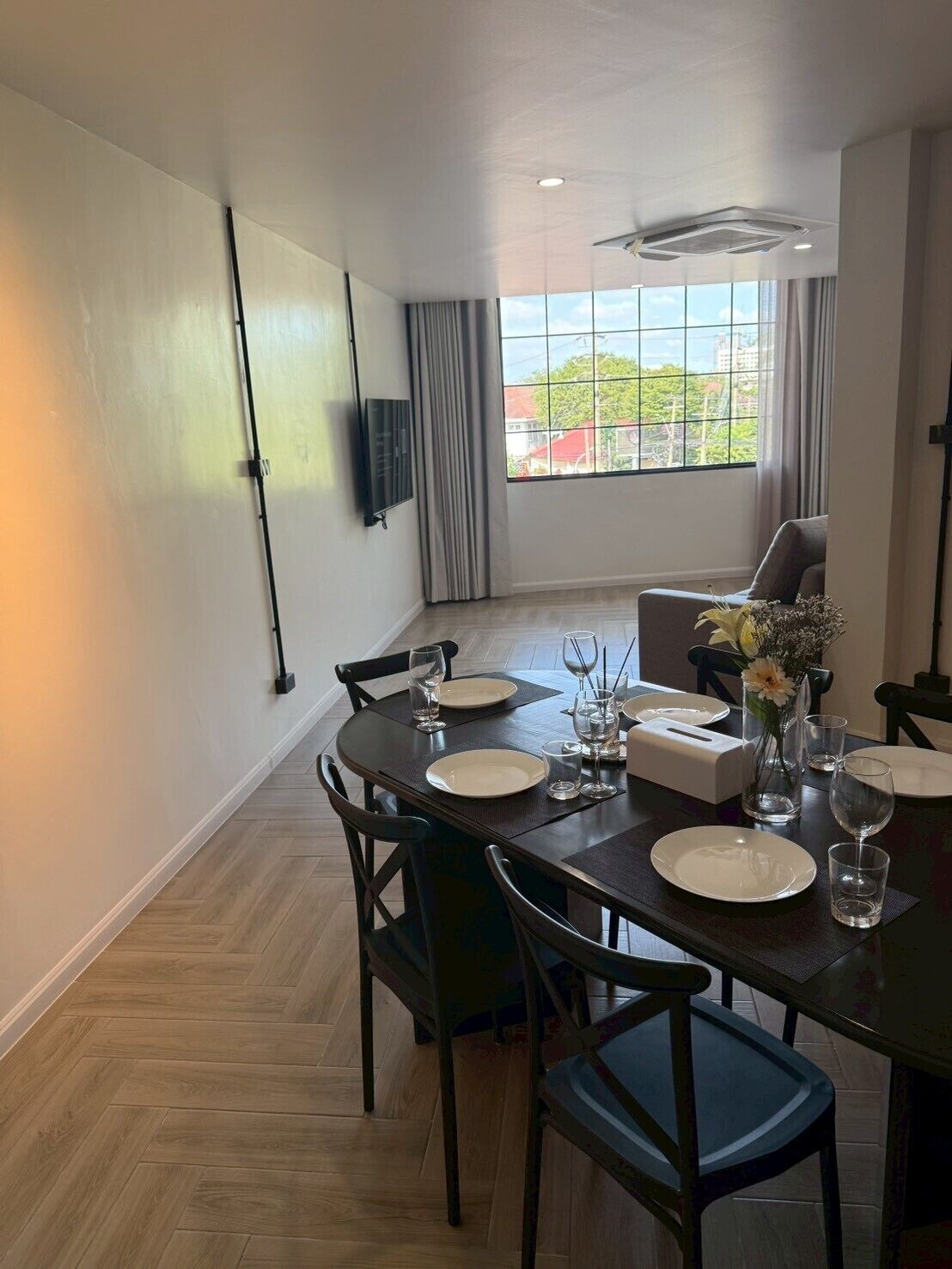 A dining area with a round table set for four, a flat-screen TV mounted on the wall, and a window with curtains.