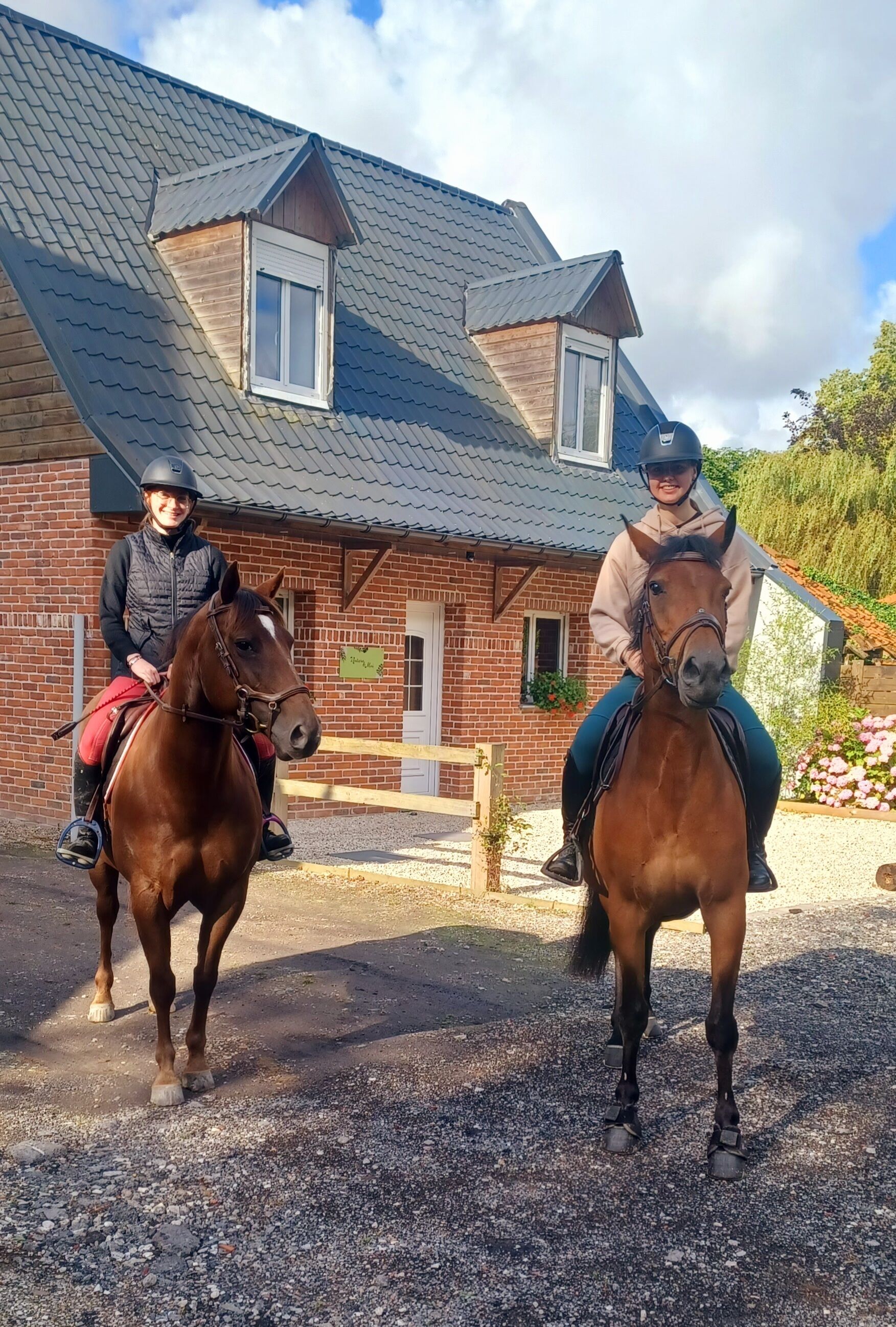 Two people riding horses in front of a brick building with a sloped roof.