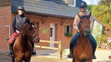 Two people riding horses in front of a brick building with a sloped roof.