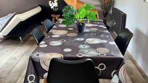 A dining area with a table set for four, surrounded by black chairs. A large potted plant is on the table, and there is a staircase in the background. - Whole House (Wittes)