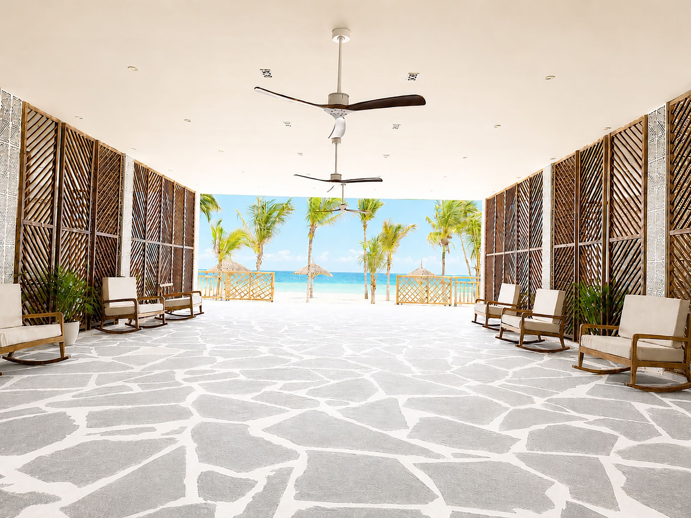 Amplia habitación con suelo de piedra estampada, ventilador de techo y vista a la playa con palmeras.