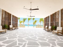 Amplia habitación con suelo de piedra estampada, ventilador de techo y vista a la playa con palmeras.
