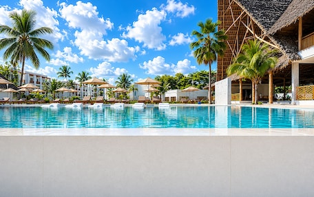A resort pool with lounge chairs, palm trees, and a building with a thatched roof.