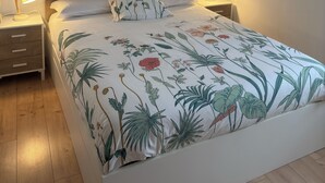 Ein ordentlich eingerichtetes Schlafzimmer mit einem hölzernen Kopfende, einer blumigen Bettdecke, Nachttischlampen und gerahmten Bildern an der Wand.
