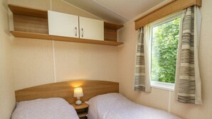 Ein kleiner, gemütlicher Raum mit zwei Betten, einem Nachttisch mit Lampe, einem Holzregal über den Betten und einem Fenster mit Vorhängen.