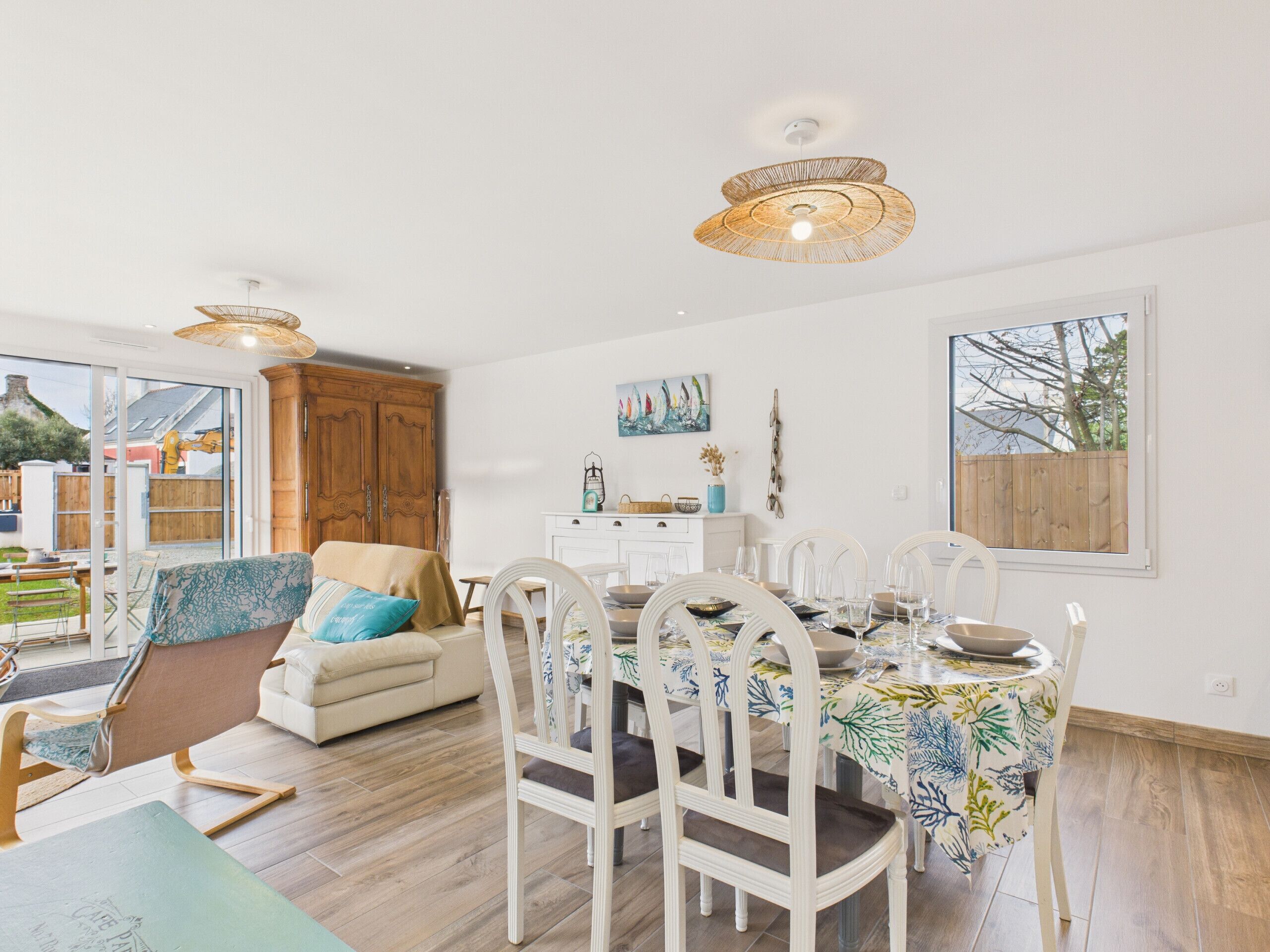 Amplia sala de estar con una mesa de comedor lista para cuatro personas, rodeada de sillas blancas. También se ve un sofá y un mueble de madera.