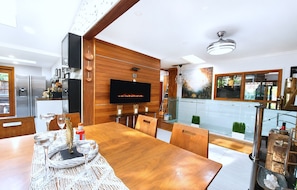 A modern kitchen with a flat-screen TV, wooden cabinetry, and a dining area with a wooden table and chairs. - Famous Palm Beach Home! 120-jet Spa Cabana, Games Room & Prime Central Location! (Sydney)