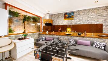 A modern living room with a foosball table, a sofa with patterned cushions, a kitchen area with a stone backsplash, and a large window with a view of greenery.