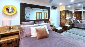 Un dormitorio moderno con una cama grande, varios almohadones, una mesita de noche con lámpara, un espejo redondo y un ventilador de techo.