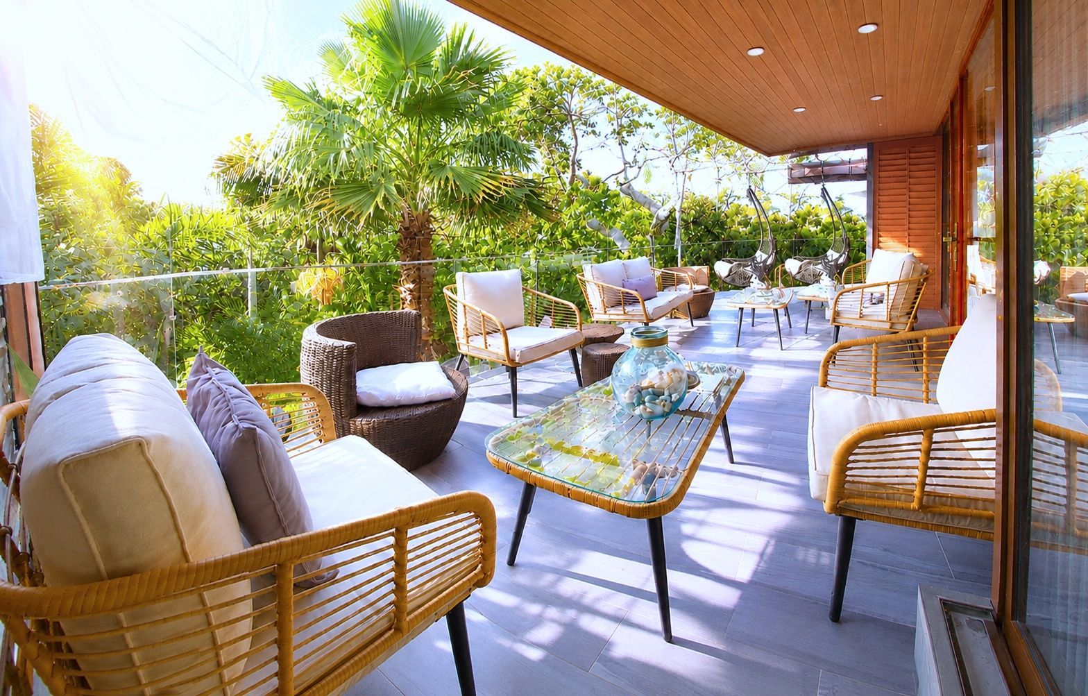 An outdoor patio with wicker furniture, a glass-top table, and a view of lush greenery.