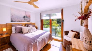 A bedroom with a large bed, a ceiling fan, a nightstand, a bedside table, a window with a view of greenery, and a decorative vase.