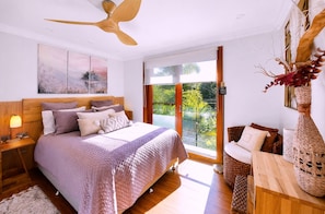 A bedroom with a large bed, a ceiling fan, a nightstand, a bedside table, a window with a view of greenery, and a decorative vase. - Famous Palm Beach Home! 120-jet Spa Cabana, Games Room & Prime Central Location! (Sydney)