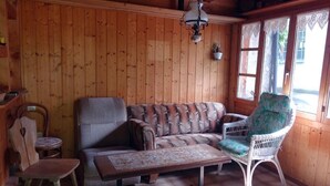 Ein gemütliches Zimmer mit Holzwänden, einem gemusterten Sofa, einem Korbstuhl, einem kleinen Tisch und einer hängenden Leuchte.