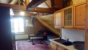 Ein gemütliches Schlafzimmer im Dachgeschoss mit schrägen Wänden, Holzbalken, einem kleinen Sofa, einem gemusterten Teppich und einem Fenster, das natürliches Licht hereinlässt.