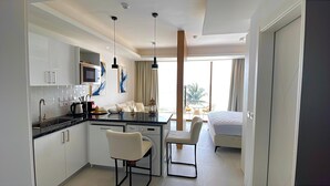 Ein modernes Hotelzimmer mit Küchenbereich, einem Esstisch mit Stühlen, einem Bett und einem großen Fenster mit Vorhängen.