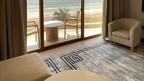 Ein Hotelzimmer mit einem großen Fenster und Blick auf den Strand und den Ozean. Es gibt einen runden Tisch mit einer Fernbedienung, einen beigen Sessel und einen gemusterten Teppich.