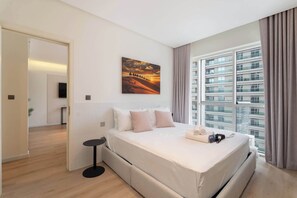 Ein modernes Hotelzimmer mit einem großen Bett, einem Flachbildfernseher, einem Sonnenuntergangsbild und Blick auf die Stadt durch große Fenster.
