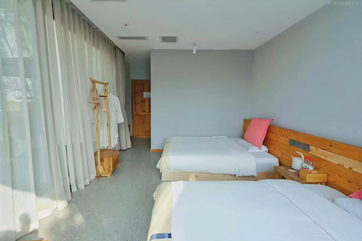 A hotel room with two beds, a wooden headboard, a bedside table with a lamp, and a clothing rack with hanging clothes.