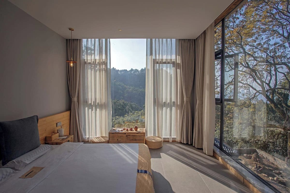A bedroom with a large bed, wooden headboard, and a view of a forest through floor-to-ceiling windows.
