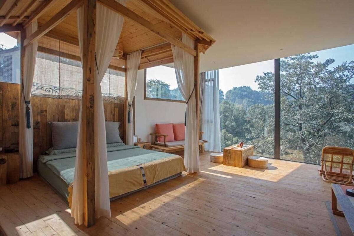 A wooden cabin bedroom with a large bed, a sitting area with a bench and cushions, and a view of trees through large windows.