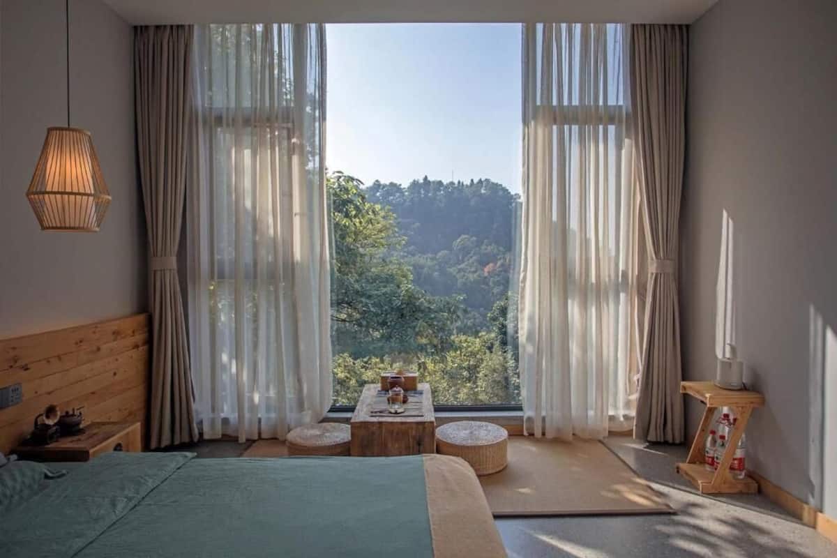 A bedroom with a large window, wooden headboard, a bed, a small table with items, and a hanging light fixture.