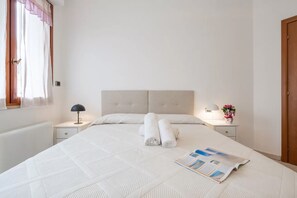 A neatly made bed with rolled towels, a bedside lamp, and a small table with a vase of flowers. - Spacious Apartment in Alghero (Alghero)