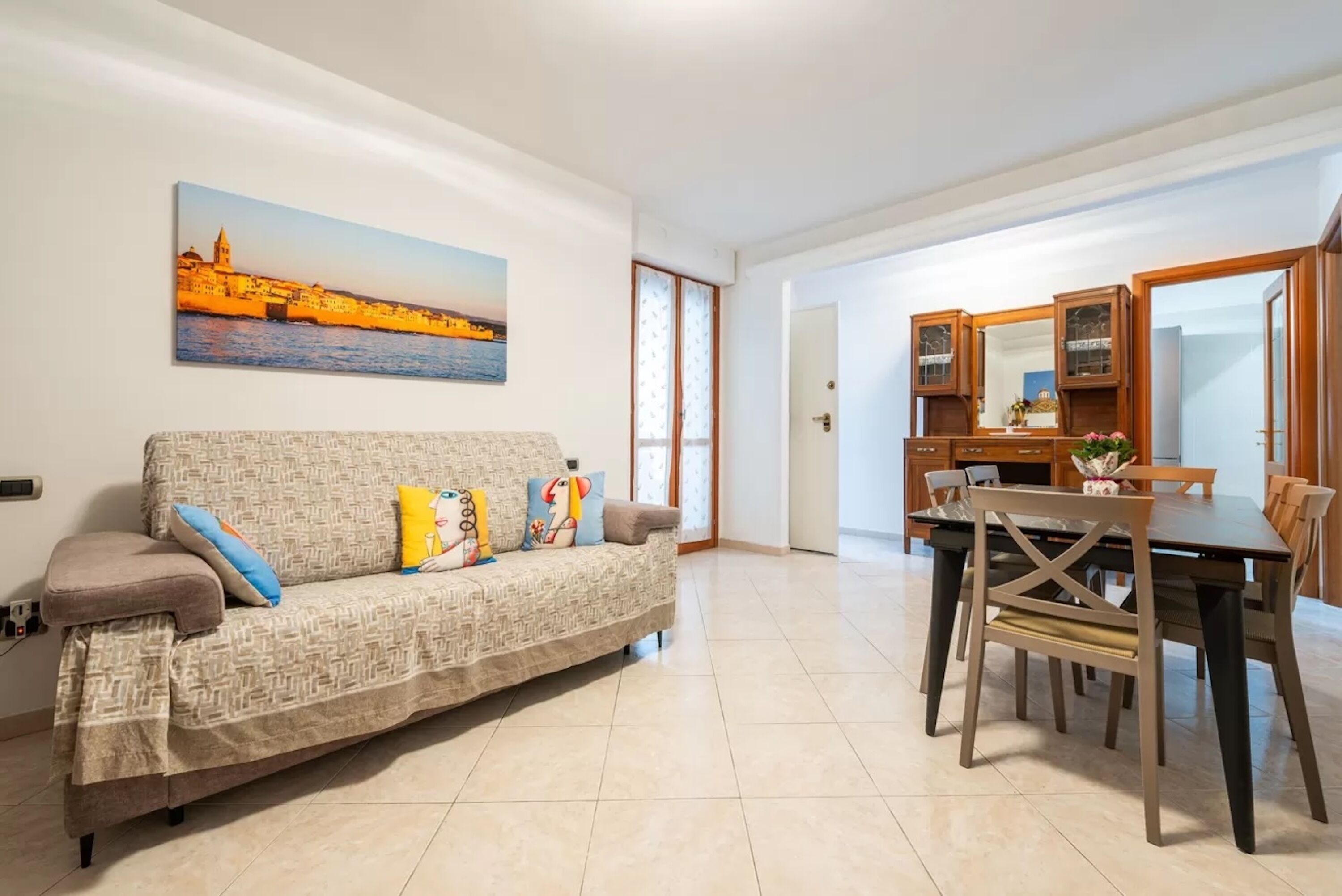 Uma sala de estar com sofá, uma mesa de jantar com cadeiras, uma cozinha e um quadro grande de uma paisagem costeira.