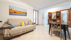 Ein Wohnzimmer mit einer Couch, einem Esstisch mit Stühlen, einer Küchenzeile und einem großen Gemälde einer Küstenstadt.