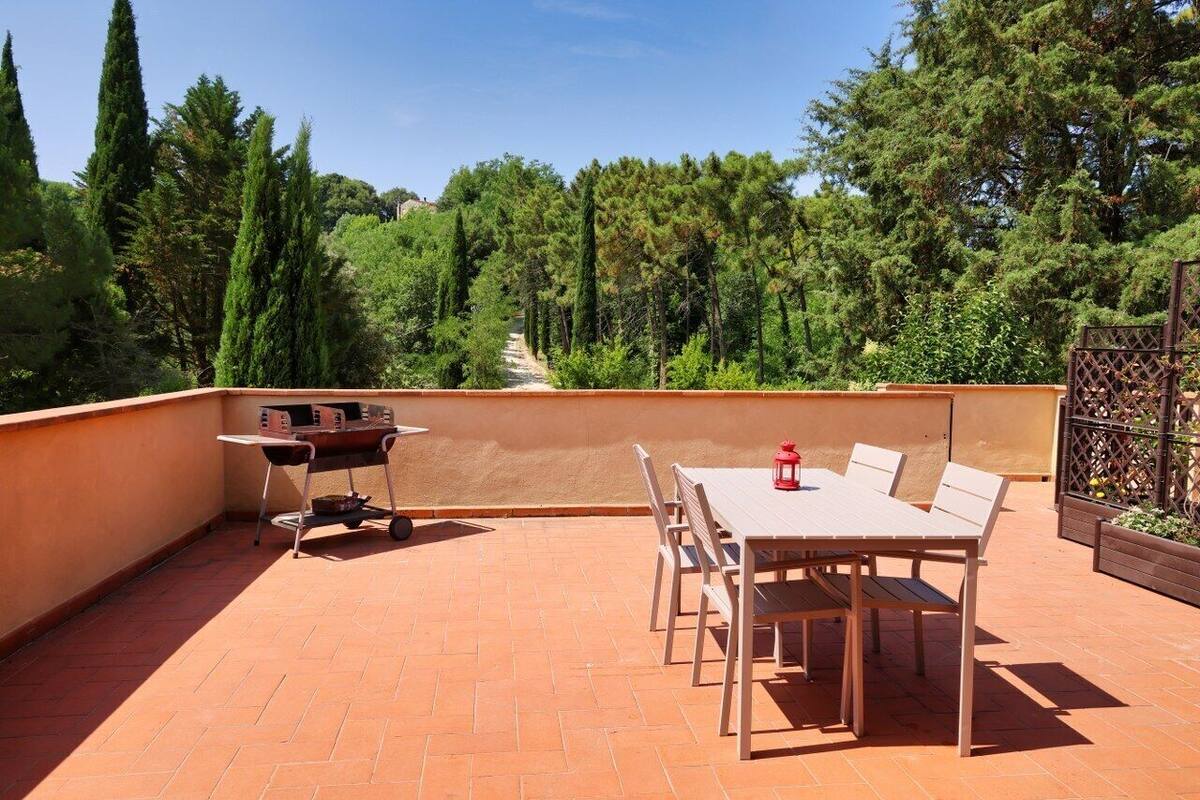 A rooftop terrace with a table and chairs, a grill, and a view of trees.