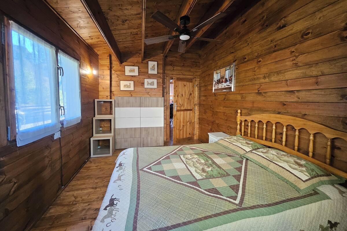 A wooden cabin bedroom with a bed, a ceiling fan, and a window with curtains.