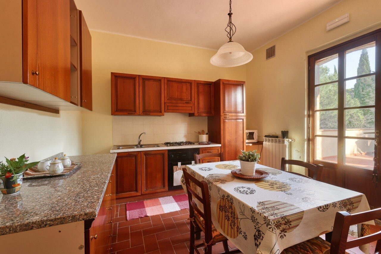 A kitchen with wooden cabinets, a granite countertop, a dining table with a tablecloth, and a window with a view of trees.