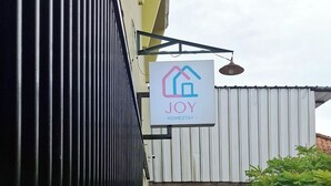 Letrero que dice 'JOY Homestay' colgado en un edificio con fachada de metal corrugado y cerca de metal negro.