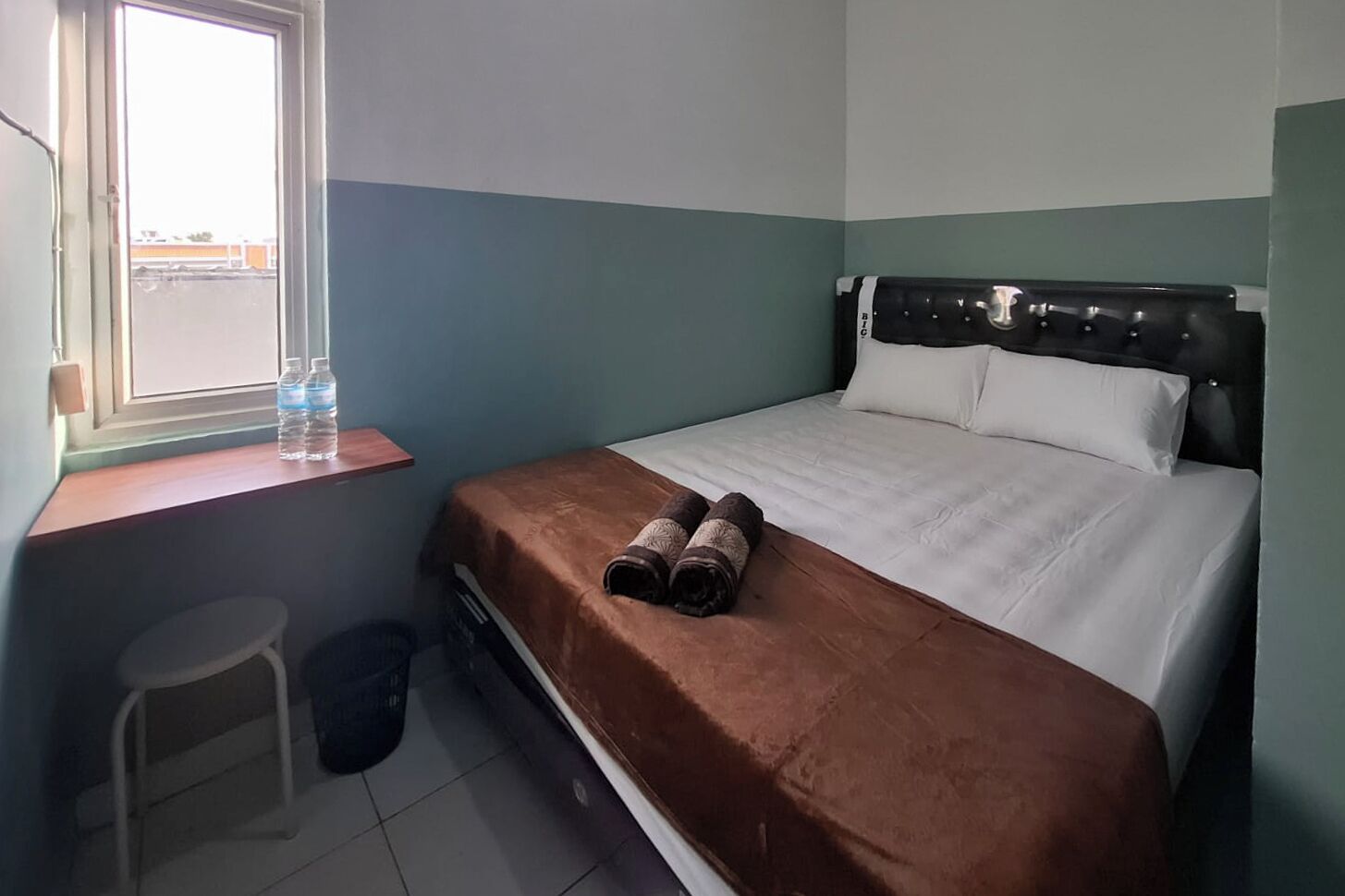 A small, clean room with a bed, a window, a stool, and a trash bin.