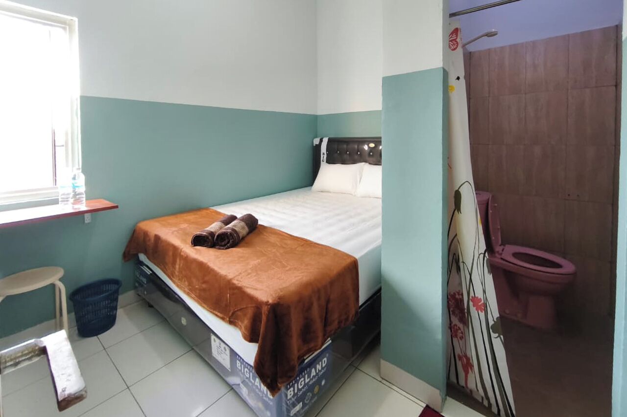Een kleine kamer met een bed, een raam, een kruk en een toilet.