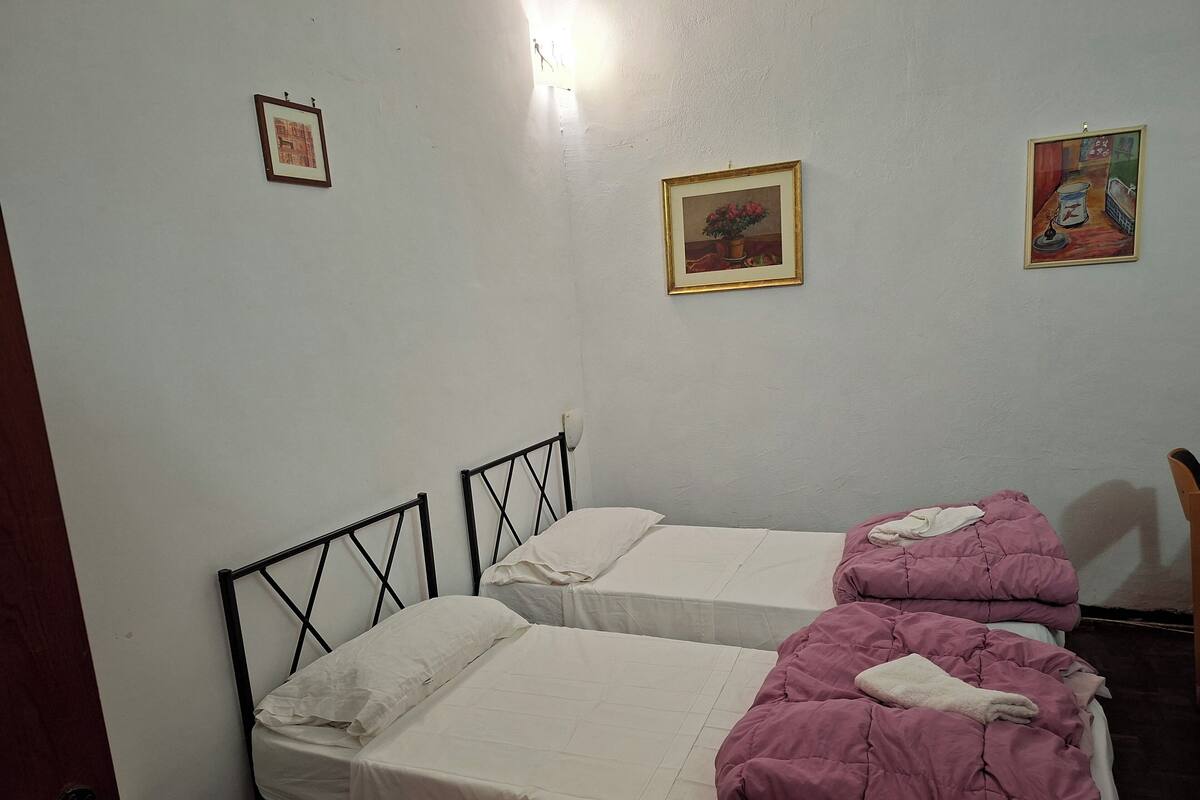 A room with two beds, each with a headboard and a white mattress. There are two framed pictures on the wall, one depicting a building and the other a still life.