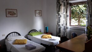 A room with two single beds, a wooden table, a blue lamp, and a window with a view of greenery. - Bed in Siena (Siena)