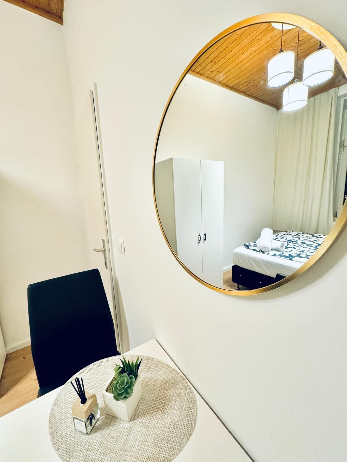 Una habitación con un espejo redondo, una cama con colcha estampada, una mesita con planta y un candelabro, y una silla.