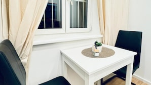 Ein kleiner Raum mit einem weißen Tisch, zwei schwarzen Stühlen, einem fenster mit Vorhängen und einem Blumentopf auf dem Tisch.