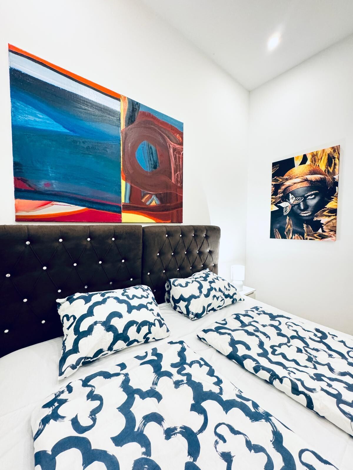 Une chambre à coucher moderne avec une tête de lit capitonnée, une literie à motifs et deux tableaux abstraits accrochés au mur.