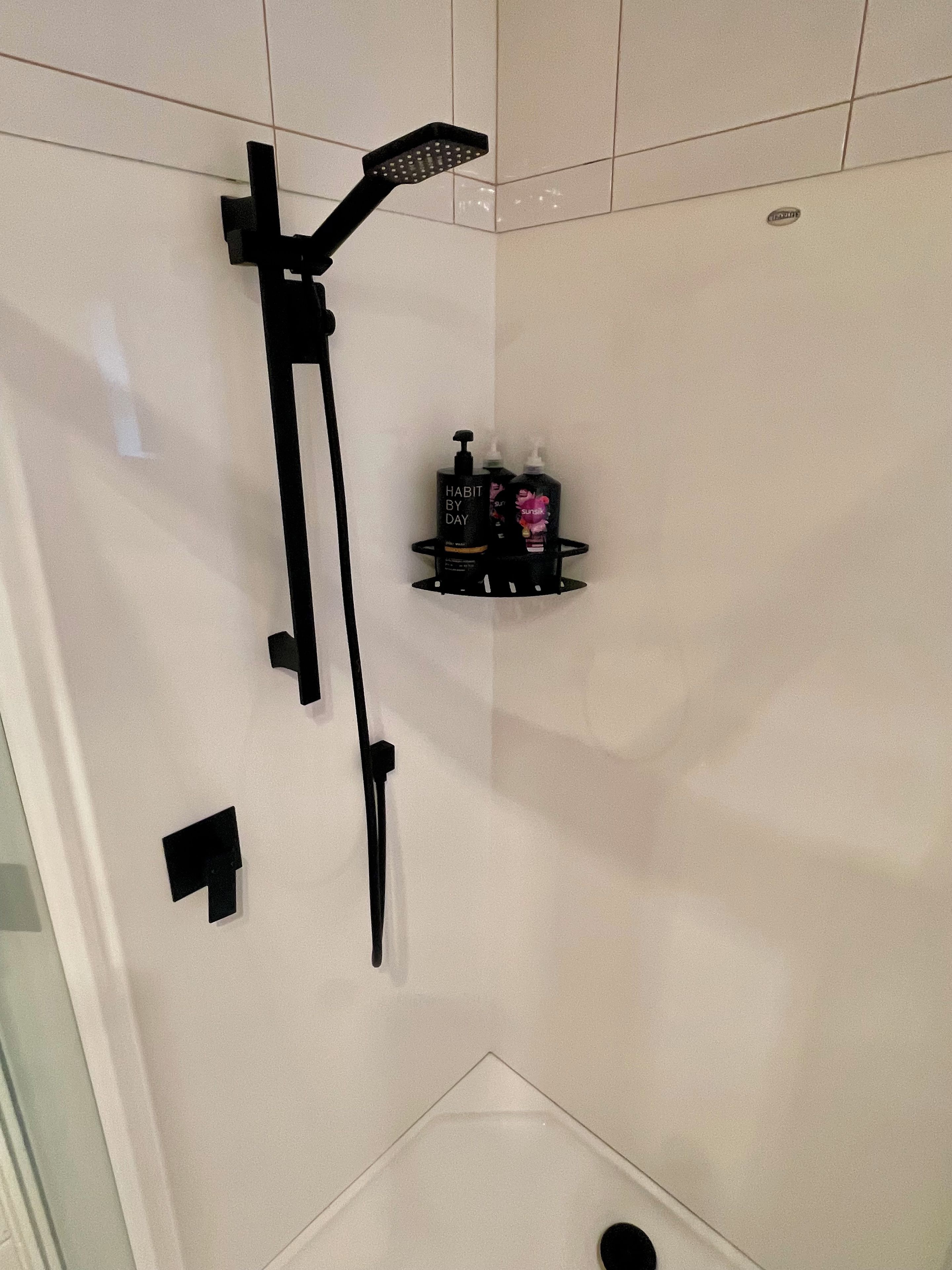 A shower with a handheld showerhead and a wall-mounted control panel. There is a black shelf with two bottles of shampoo and conditioner.