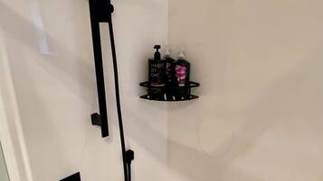 A shower with a handheld showerhead and a wall-mounted control panel. There is a black shelf with two bottles of shampoo and conditioner.
