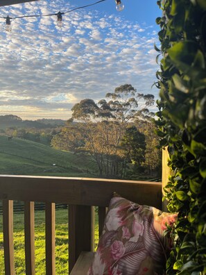 A balcony with a view of a hilly landscape, a wooden railing, and a floral pillow. - Swallows Nest Cottage -self contained with amazing views and outdoor bath. (Waiatarua)