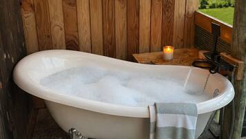 A wooden cabin bathroom with a freestanding bathtub, string lights, and a view of the countryside.