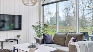 Een moderne woonkamer met een flatscreen televisie, een ronde salontafel met glazen en een boek, een grijze bank met kussens en een groot raam met uitzicht op een tuin.