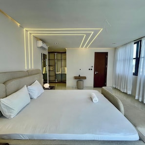 A modern hotel room with a large bed, a bedside table, a mirror, a vanity with a sink, and a view of the city through large windows. - Deluxe Haven Apartment (Lekki)
