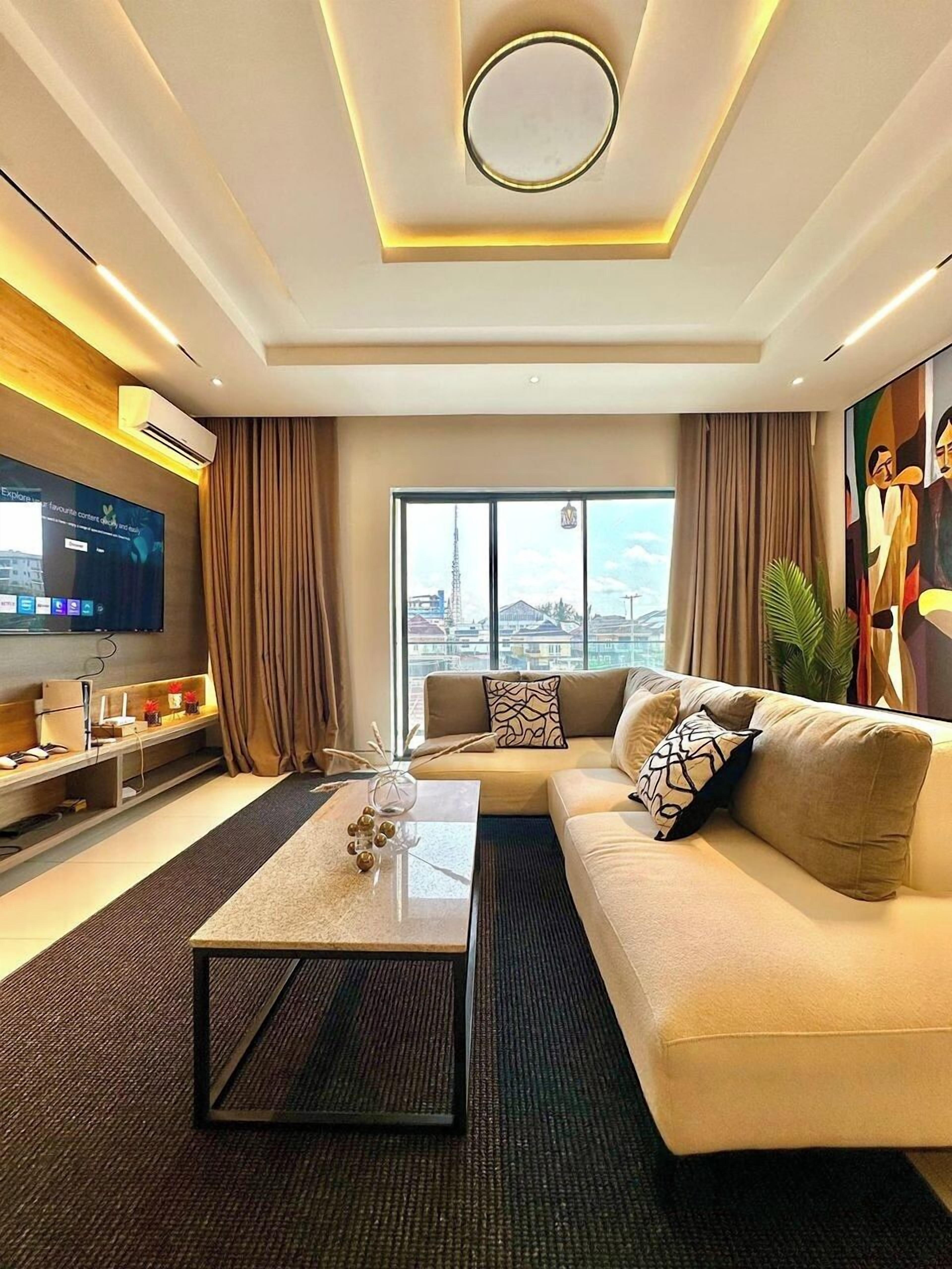 A modern living room with a large flat-screen TV, a sofa, a coffee table, and a view of the city through large windows.