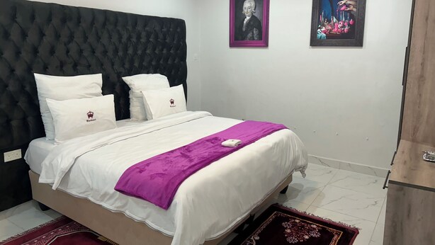 A bedroom with a tufted headboard, a bed with white bedding and a purple throw, two framed pictures on the wall, and a wooden cabinet. - Boutique Ensuite Room Mineheart Hotel, Masvingo (Masvingo)