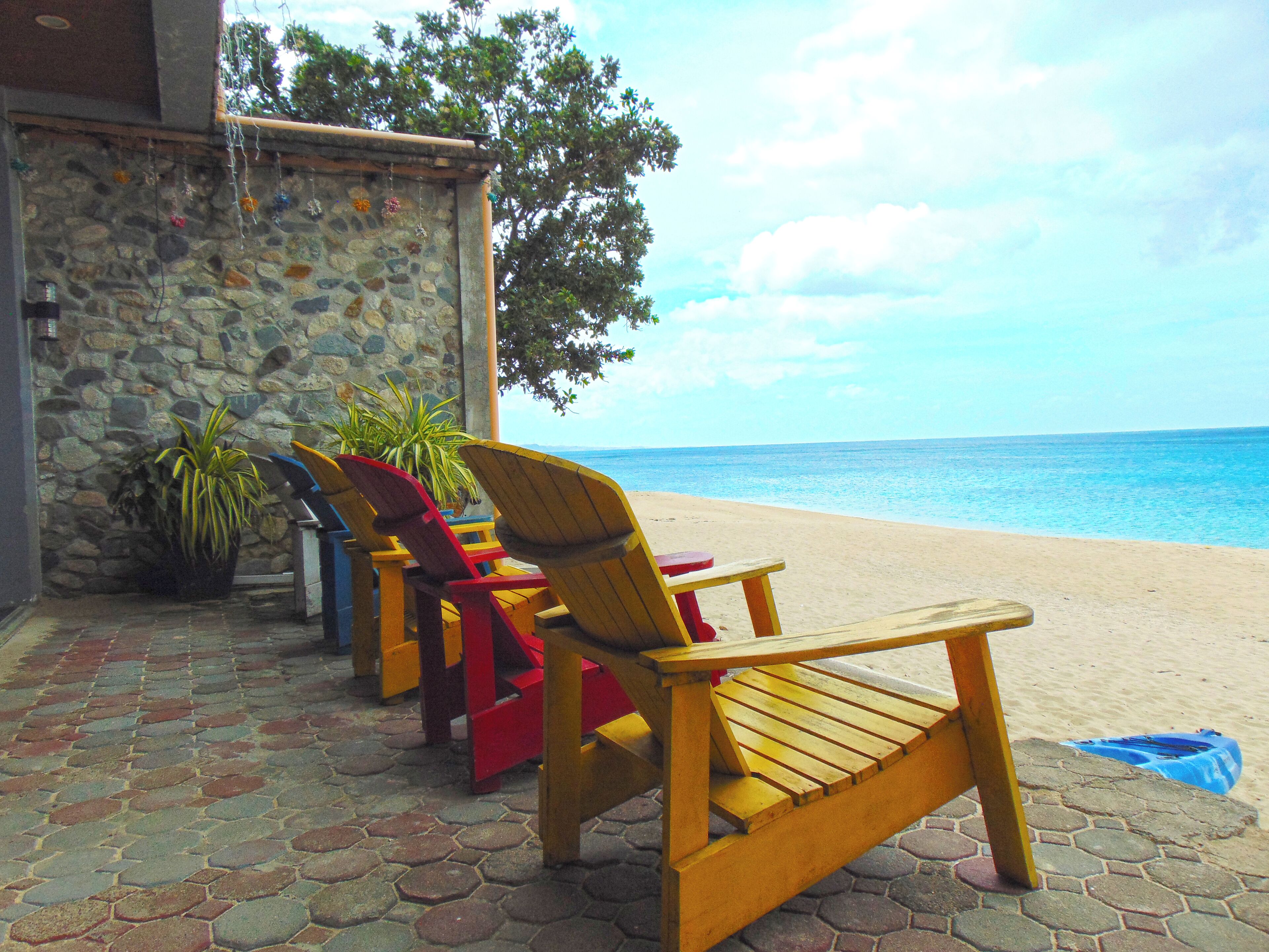 A row of wooden beach chairs facing a stone wall with a view of the ocean.