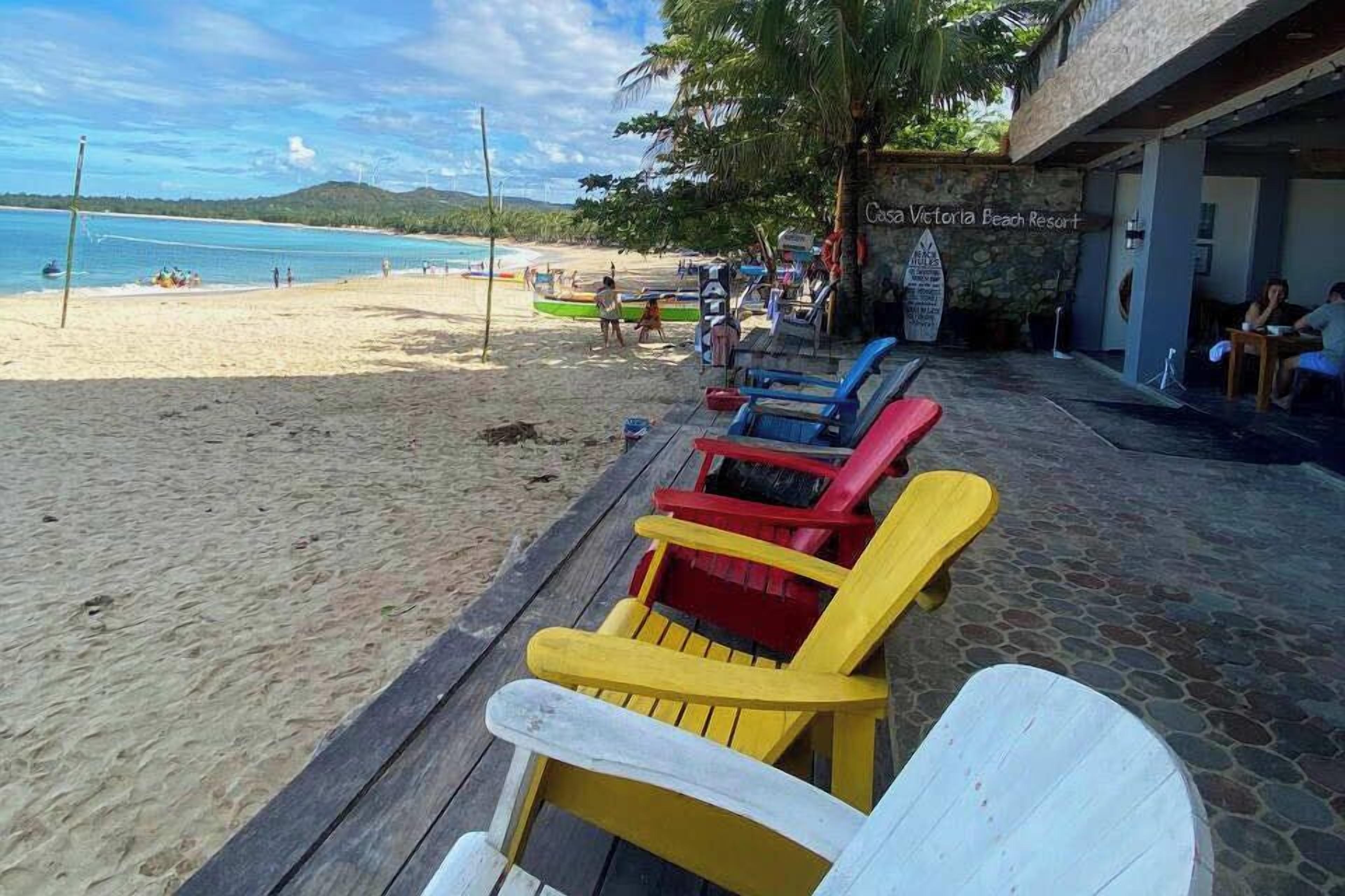 Zona d'estar a la platja amb cadires Adirondack de colors i un camí empedrat de pedra que condueix a un edifici amb un rètol que diu "Casa Victoria Beach Resort".